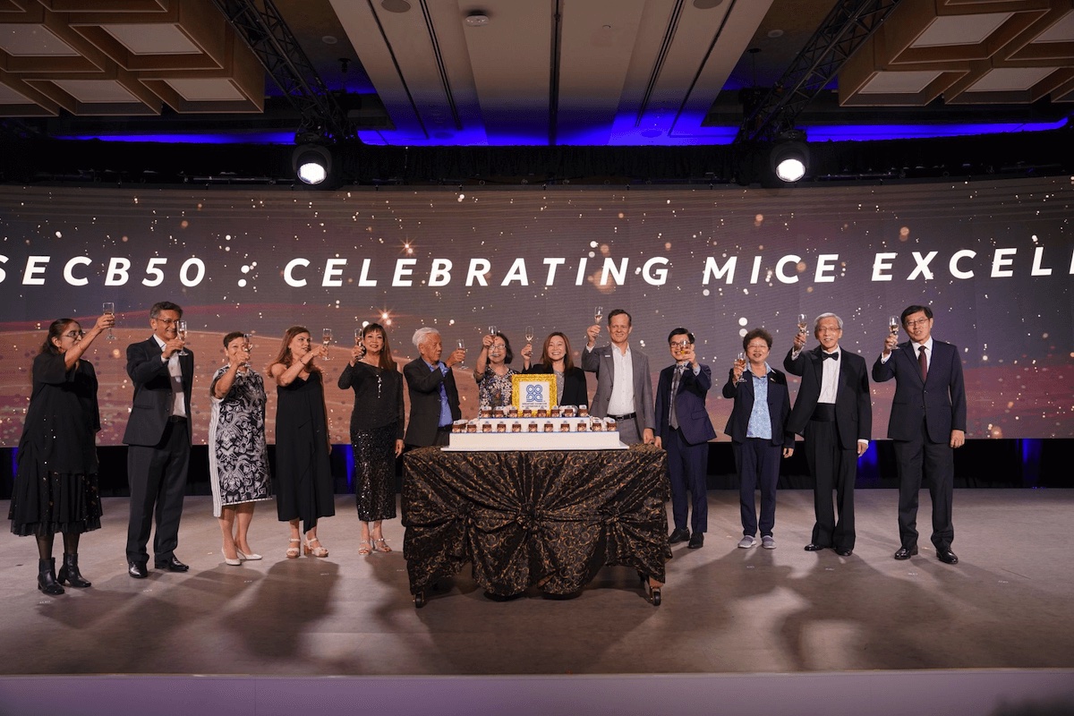 A group of people stand on a stage, posing for a photo in front of a large cake. The backdrop reads "SECB50 - Celebrating MICE Excellence." The setting appears to be a formal event.