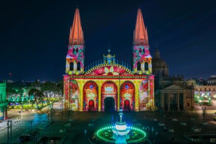 A cathedral at night displays colorful illuminated projections on its facade, with a lit fountain in the foreground and surrounding plaza nearly empty.