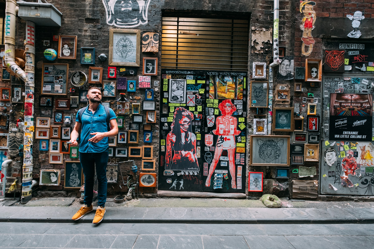 A person stands in front of a wall covered in various artworks and framed pictures in a narrow alleyway in Melbourne. They wear a blue shirt, jeans, and tan boots, with a backpack slung over their shoulders.