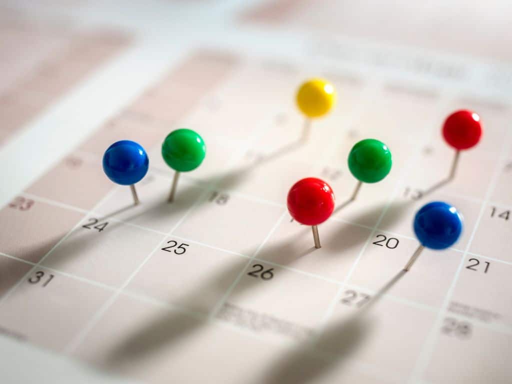 Close-up of a calendar with various pushpins marking different dates, hinting at event planning. A pencil lies on the open page, suggesting meticulous scheduling. The background is a blue wooden surface, adding a touch of calmness to this business plan essential.