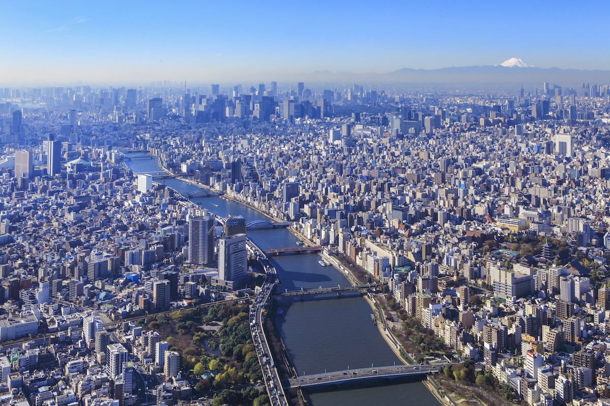 Aerial view of Tokyo's sprawling cityscape with multiple rivers and bridges, densely packed buildings, and mountains in the distant background under a clear, blue sky.