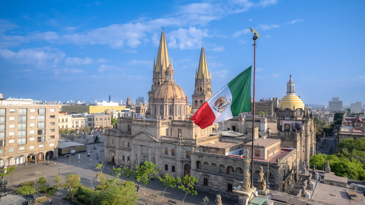 A view of Guadalajara Cathedral with its twin gold spires and the Mexican flag in the foreground, all under a clear blue sky.