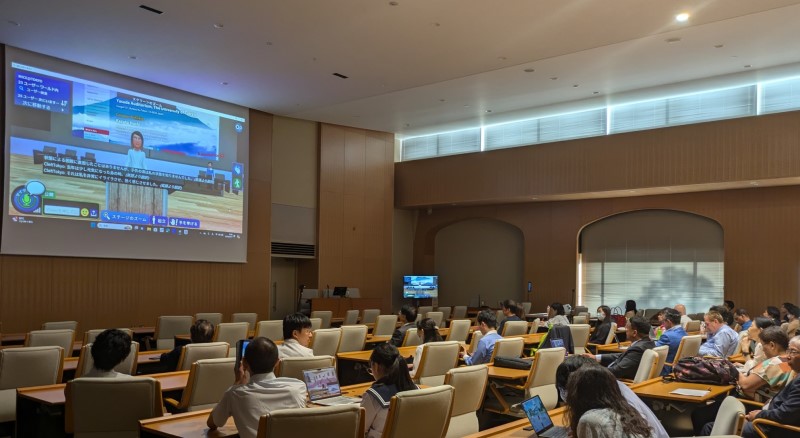 A group of people sits in a conference room with a large screen projecting a video presentation at the front.