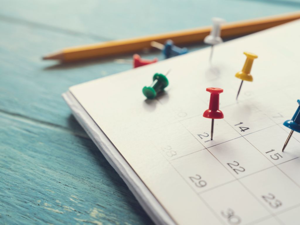 Close-up of a calendar with multiple push pins marking dates. A pencil lies next to the calendar on a wooden surface.