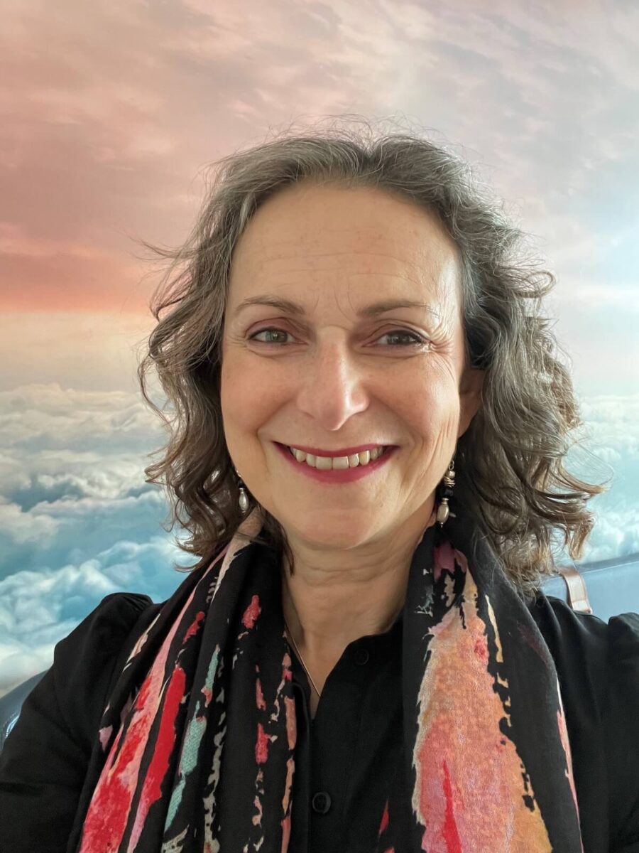 A person with curly gray hair and a colorful scarf smiles in front of a backdrop featuring clouds and a gradient sky.