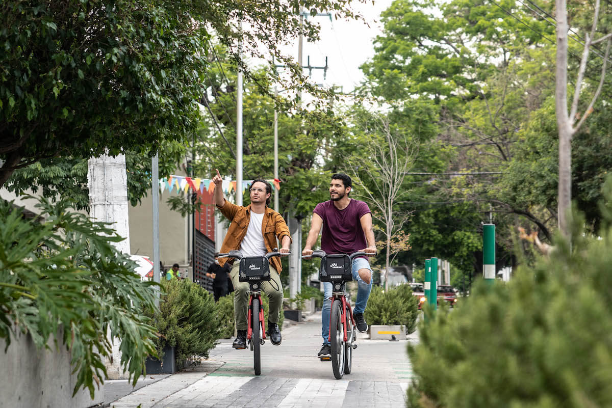 Two men ride bicycles on a tree-lined path, with one pointing ahead and both looking in the same direction. The scene is urban with greenery and colorful decorations in the background.