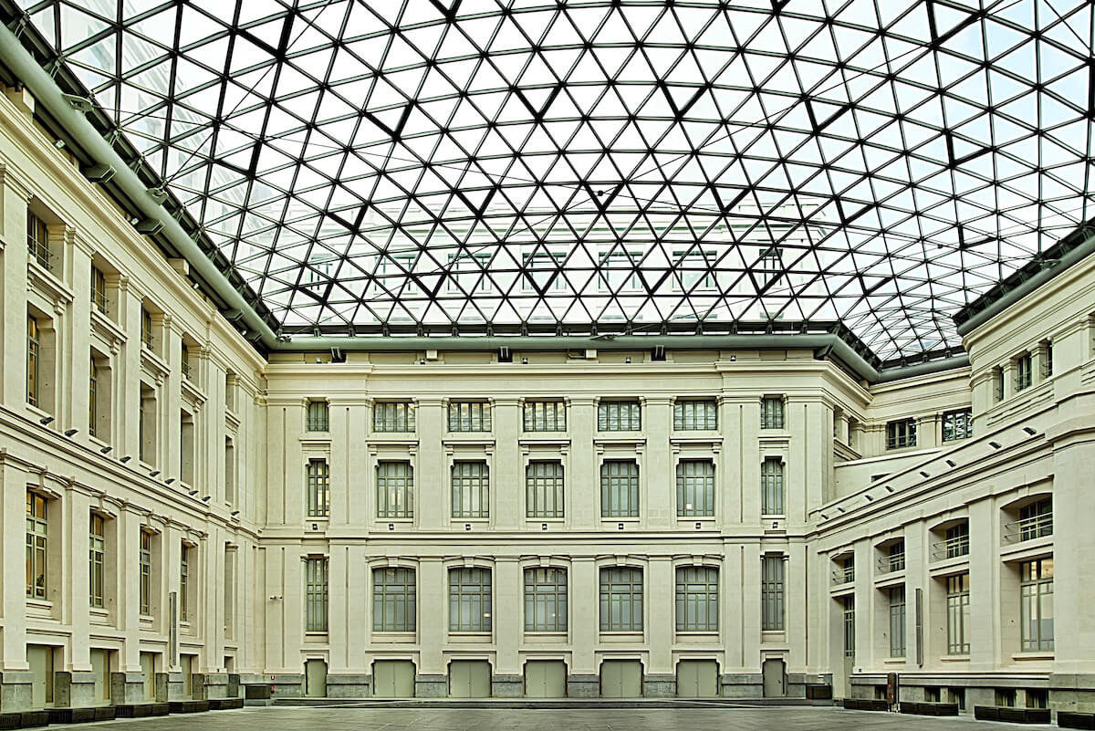 Spacious courtyard with a glass-domed ceiling, surrounded by a multi-story neoclassical building with tall windows and white columns.