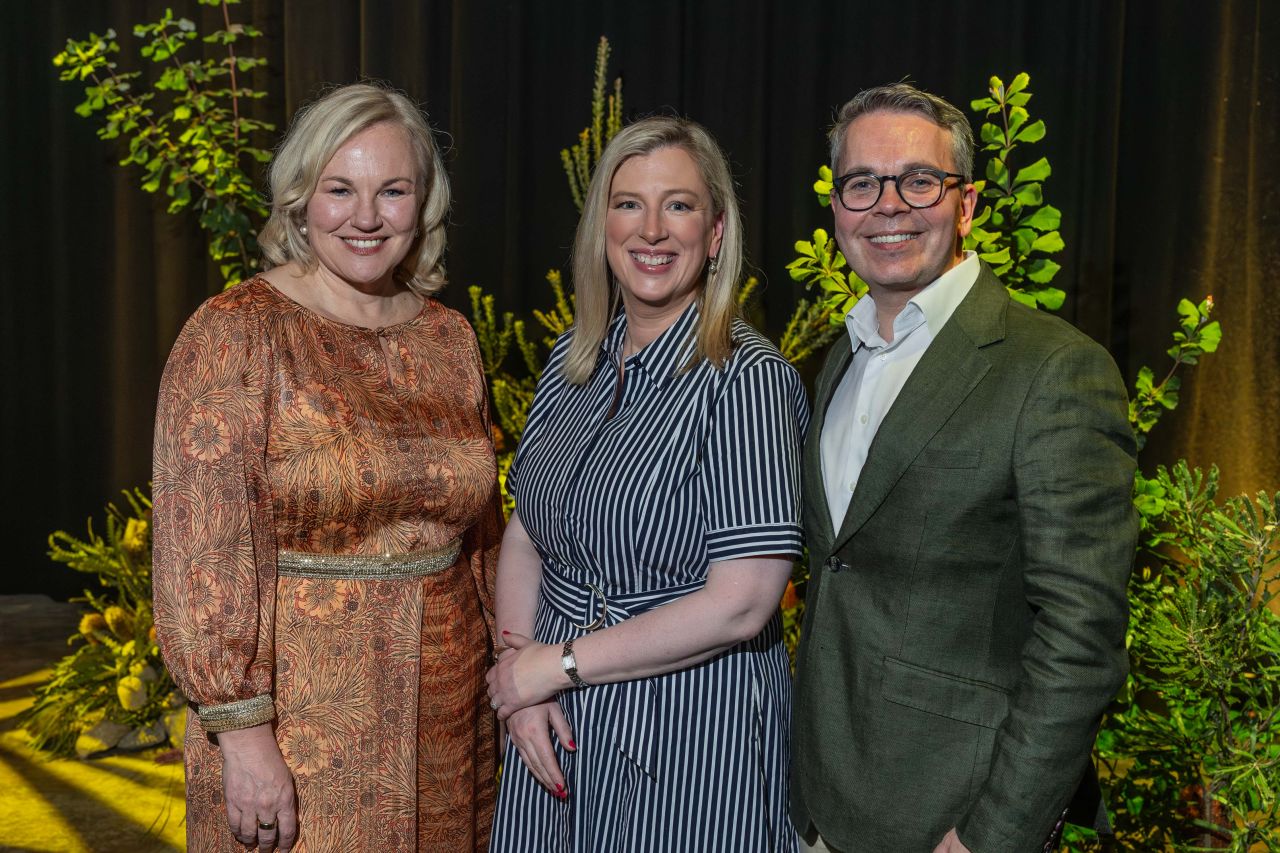 Three people stand together smiling in a garden-themed setting. The background features plants and a dark curtain.