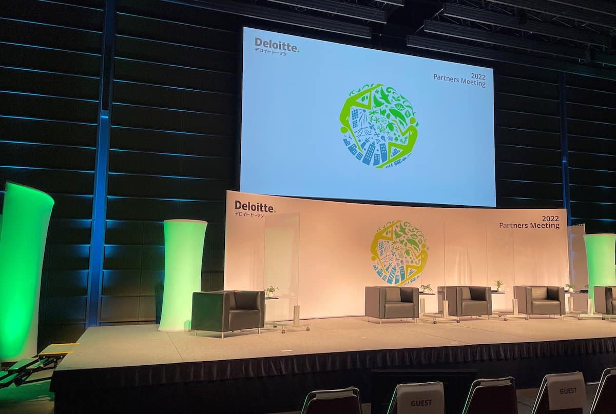 A stage setup for the 2022 Deloitte Partners Meeting, featuring a white backdrop with logos, black chairs, green lighting accents, and a projection screen displaying the event's name and logo.