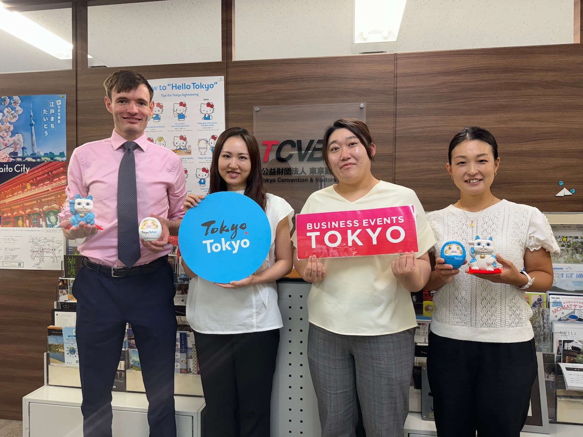 Four people stand in an office holding Tokyo tourism signs and Japanese mascot figurines. A signboard behind them reads "TCVB" and "Business Events Tokyo.