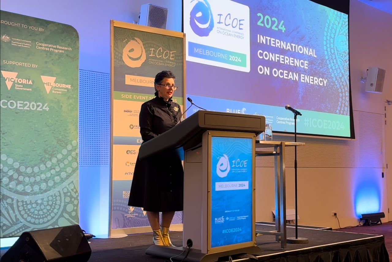 A woman stands at a podium speaking at the 2024 International Conference on Ocean Energy, with multiple screens displaying the event's title and sponsors in the background.