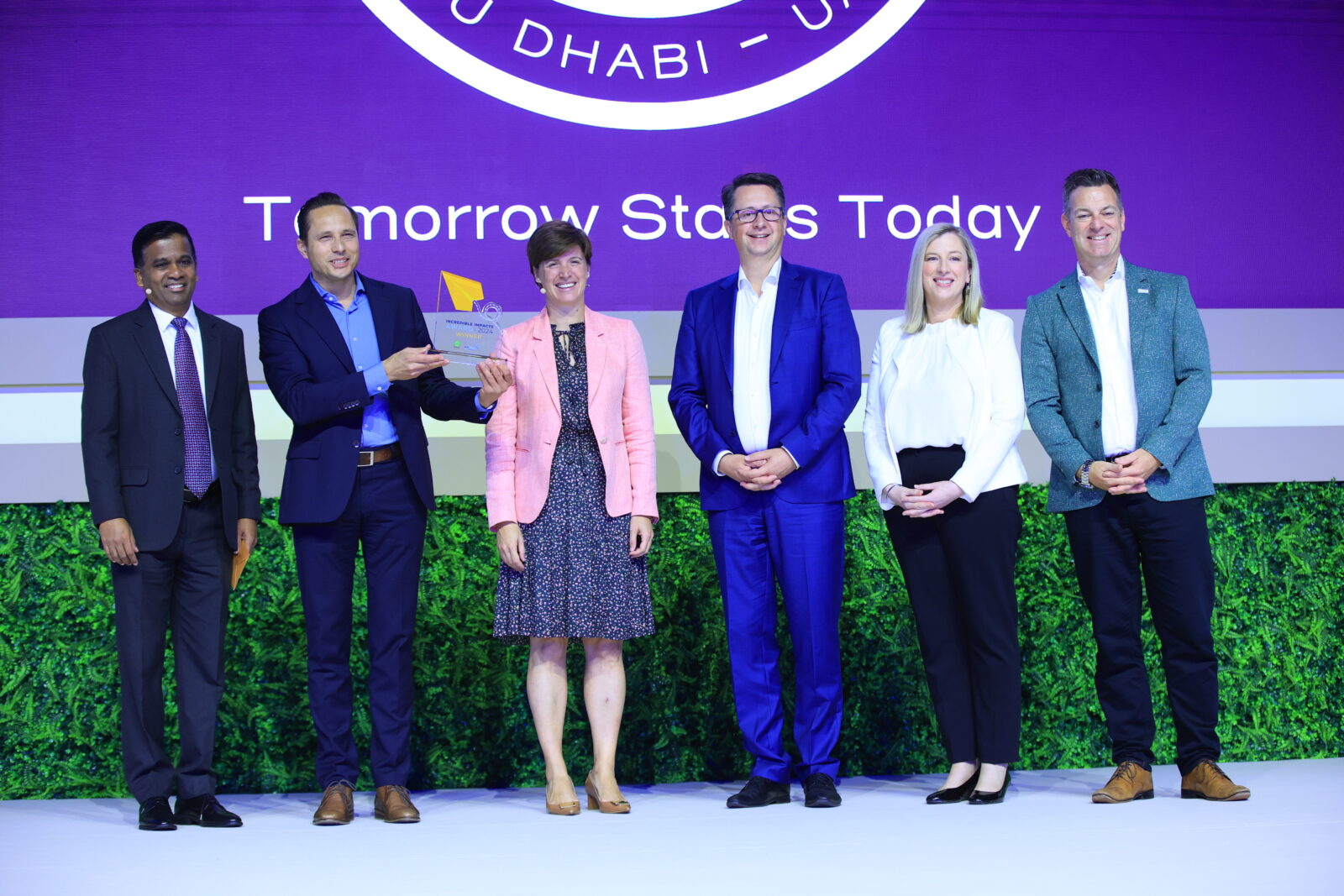 Six people stand on stage, three men and three women, posing for a group photo with one man holding a plaque. The background is purple, displaying the text "Tomorrow Starts Today.