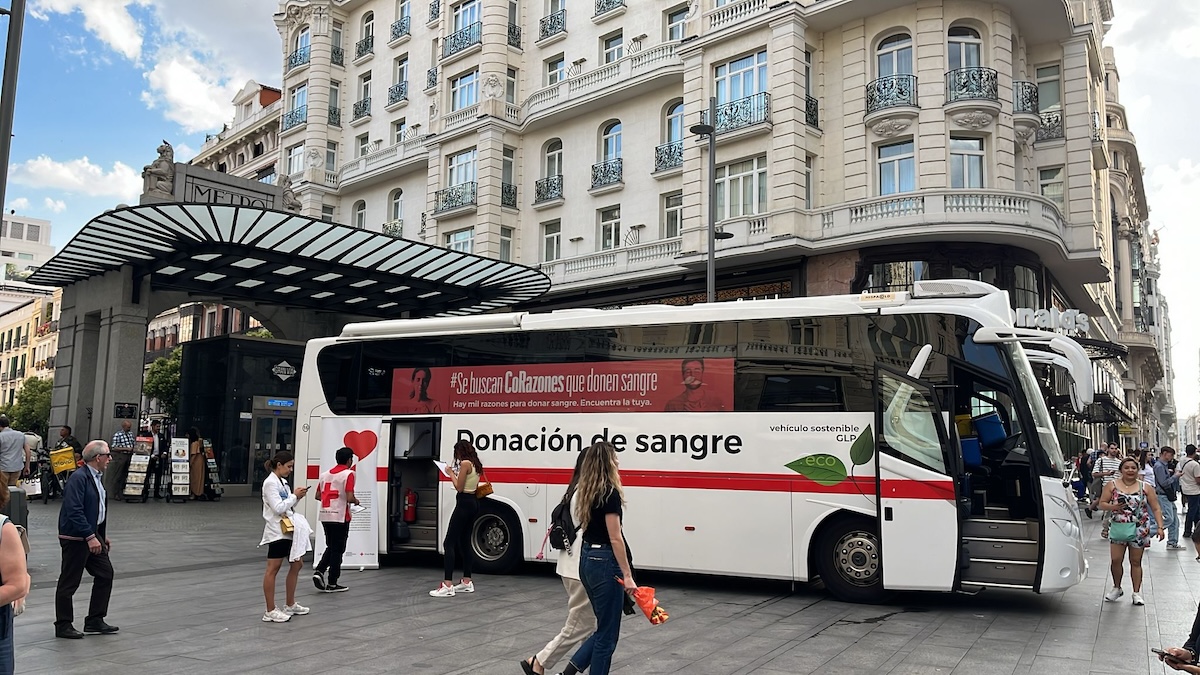 A blood donation bus is parked in a square in Madrid. People are walking by in the foreground.