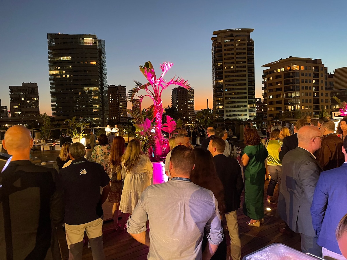 A crowd of people gathered on an outdoor patio during sunset, with pink-lit plants and tall buildings in the background.