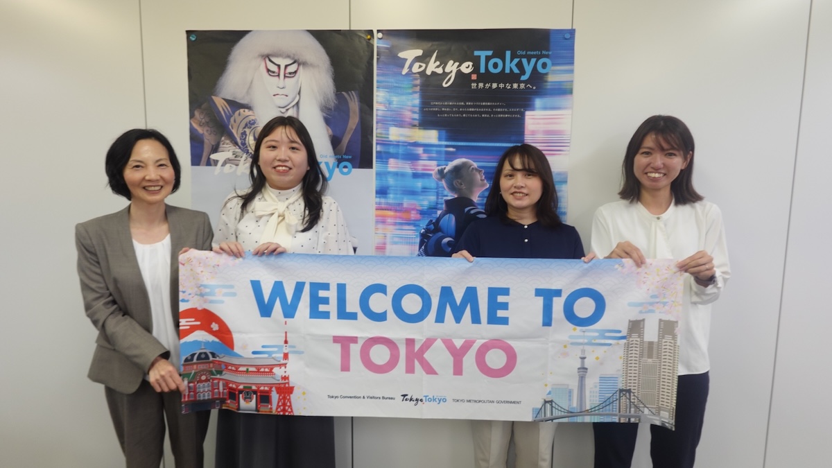 Four women stand smiling, holding a "Welcome to Tokyo" banner. Two Tokyo-themed posters are visible in the background.