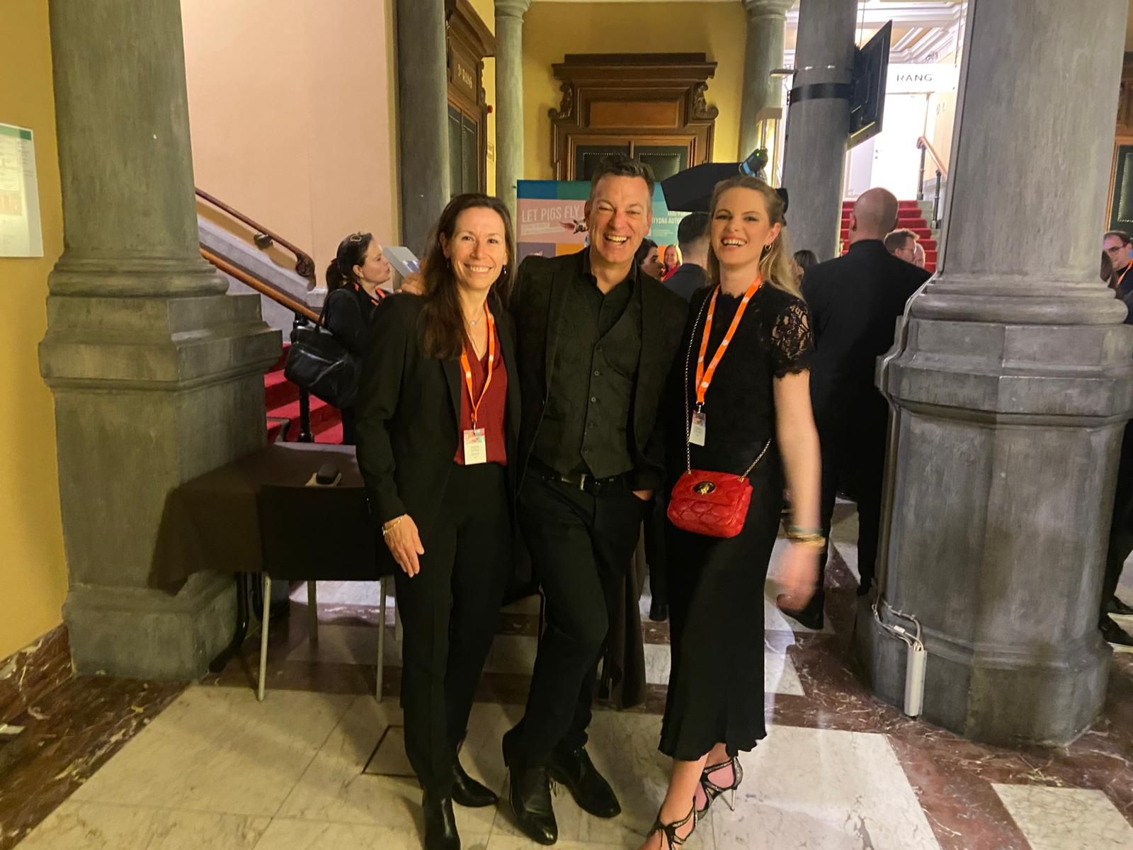 Three people smiling and posing for a photo in a building with columns and marble flooring. They are wearing semi-formal attire with event badges.