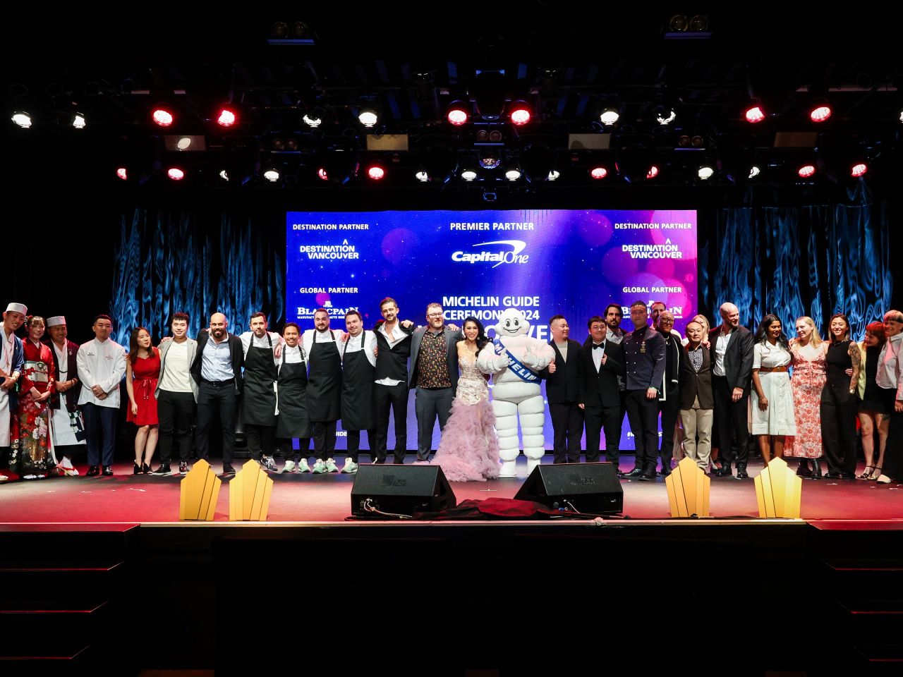 A group of people, including a person dressed as the Michelin mascot, stands on a stage in front of a banner reading "Michelin Guide Ceremony 2024.