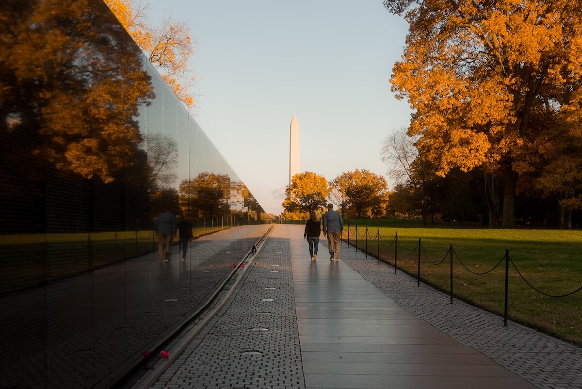 Two people walk along a reflective wall at the National Mall, with the Washington Monument visible in the distance, surrounded by autumn trees.