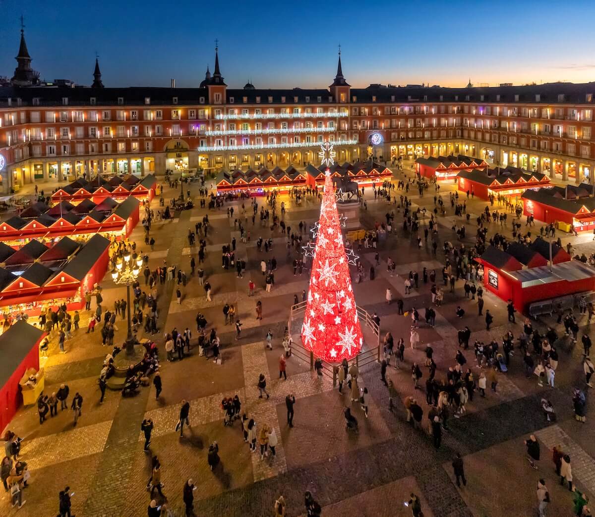 Aerial view of a festive square at dusk with a large illuminated Christmas tree and red market stalls surrounded by people.