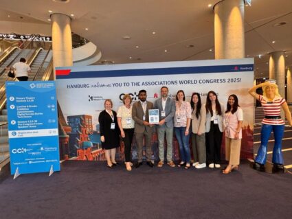 A group of eight people stands posing indoors in front of a banner for the Associations World Congress 2025 in Hamburg, next to an event schedule and a sailor statue.