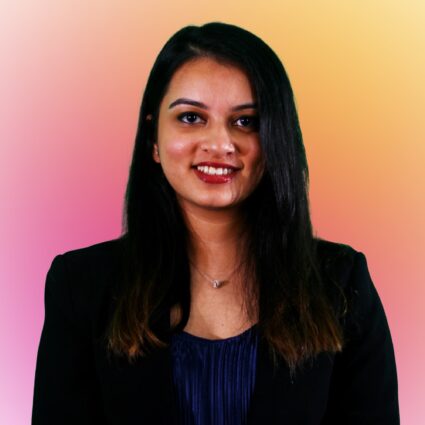 A woman with long dark hair wearing a dark blazer and blue top stands in front of a pink and yellow gradient background, smiling at the camera.