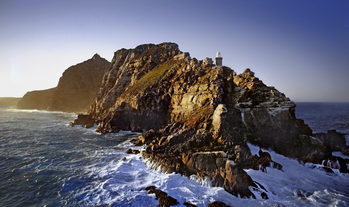 Rocky headland with a lighthouse on top, surrounded by ocean waves crashing against the cliffs under a clear sky.