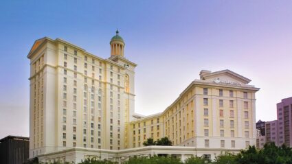 Large cream-coloured hotel building with multiple floors, tall windows, a domed tower, and the "Southern Sun" logo on the facade, set against a clear sky.